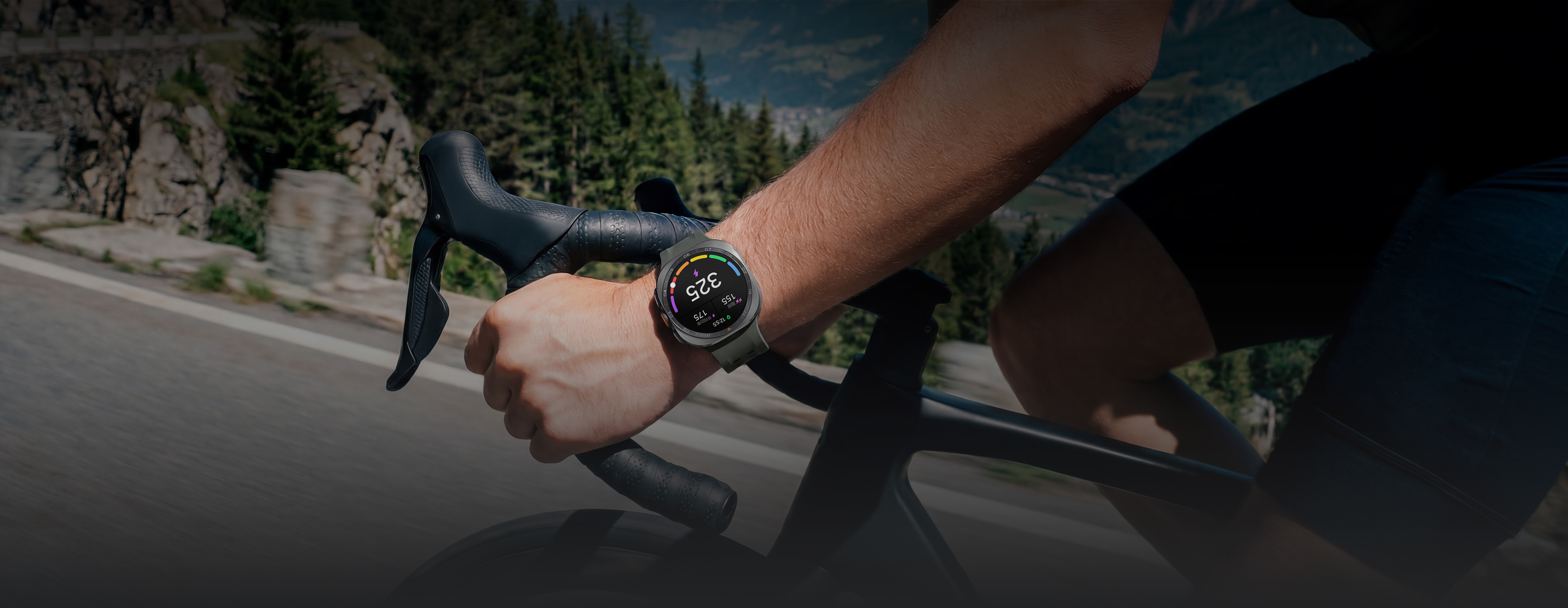 画面中，一名骑行者驰骋于山间公路，腕间佩戴一块钛霜银Galaxy Watch Ultra2，手表搭配绿色水上运动表带。表盘界面正在显示一组个性化的功能阈值功率（FTP）数值，并同步展示踏频、平均功率两项运动数据。