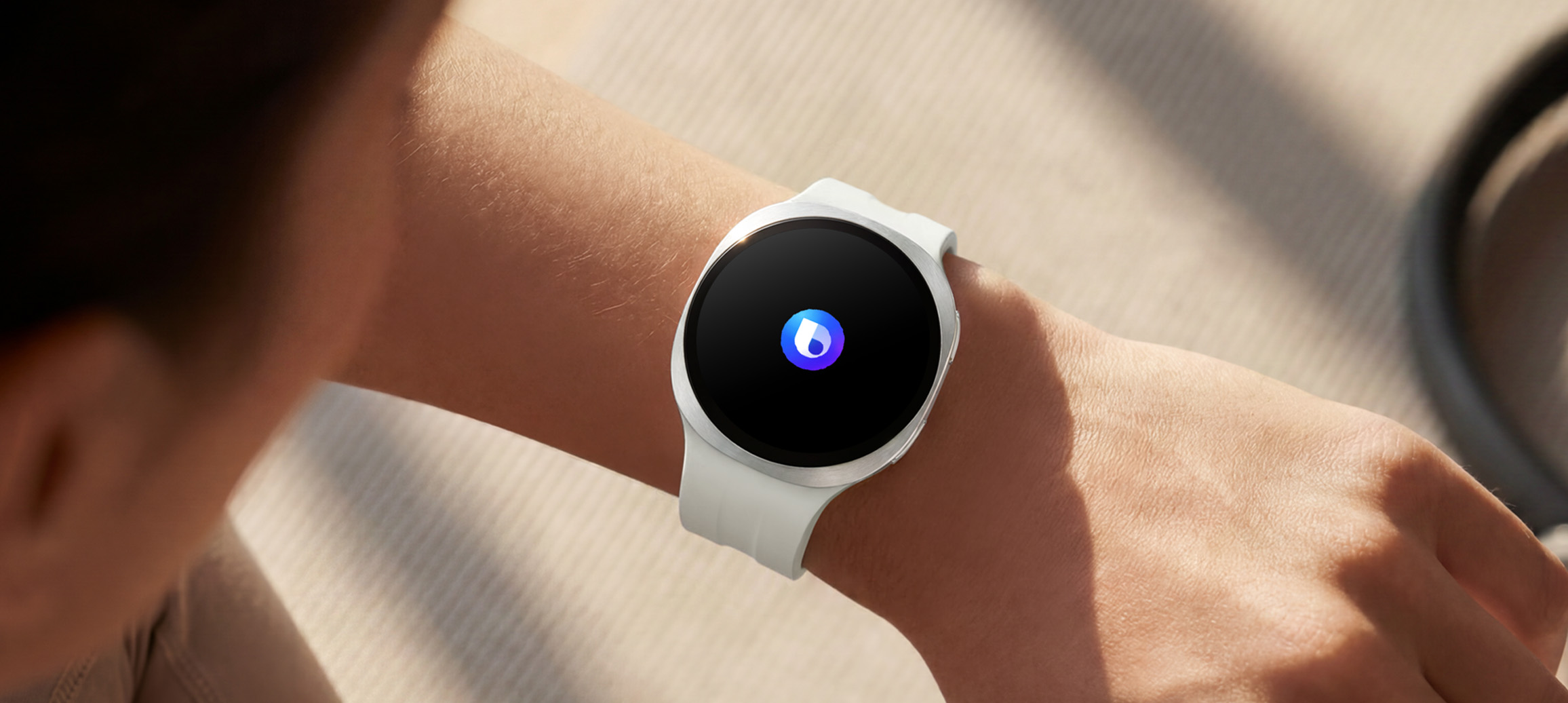 画面中的人物佩戴一块远空银色Galaxy Watch9，这款手表搭配白色运动表带。用户抬起手腕，通过Bixby向手表下达语音命令。手表屏幕显示Bixby图标，界面同步标注“询问Bixby”的字样。