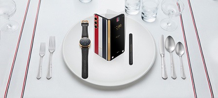 桌子上铺着白色桌布，桌布带有标志性罗缎丝带纹理封边。白色盘子上是Galaxy Watch6蓝牙版 40mm，搭配黑色卵石纹皮革表带，Galaxy Z Fold5 Thom Browne限量版，略微展开，背板玻璃的标志性罗缎饰设计和Galaxy Z Fold5 S Pen（三星折叠屏专属）。