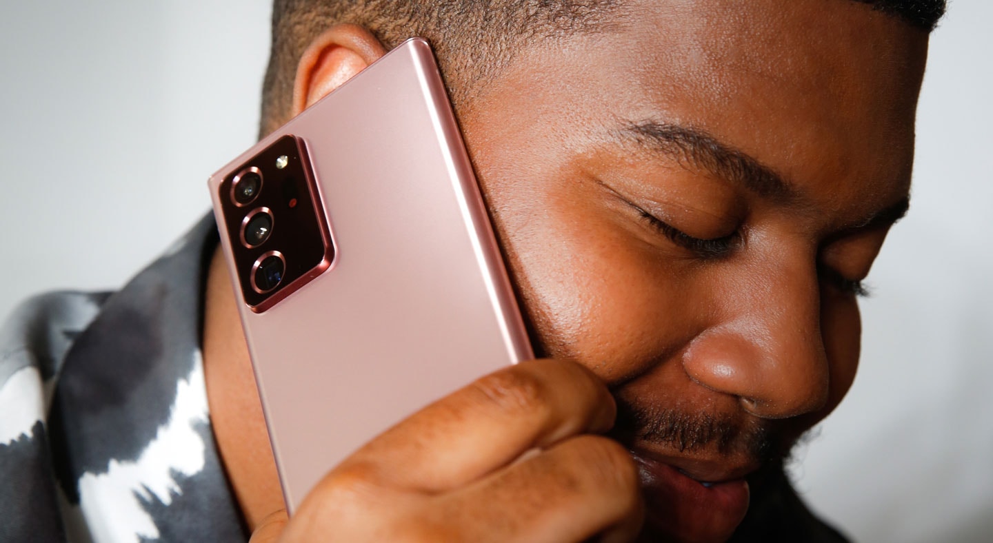 khalid手举一台曜岩黑色Galaxy Note20 5G手机贴近右耳，闭目微笑。他坐在米色的沙发上，身穿黑白双色的扎染T恤。